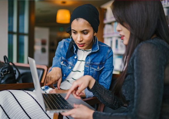 två unga kvinnor sitter tillsammans och jobbar på en laptop