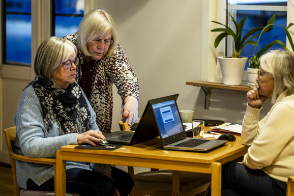 Två kvinnor vid skrivbord, de har varsin laptop, en tredje kvinna pekar mot skärmen