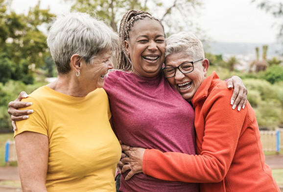 tre äldre kvinnor i färgstarka kläder kramar om varandra och skrattar hjärtligt