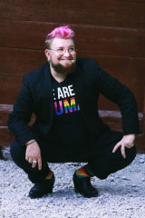 Bild på Alex sittandes på huk med klackskor och i svart kostym med en t- shirt med texten