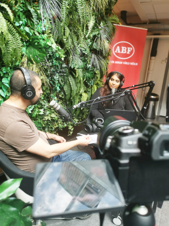 två personer sitter mitt emot varandra i en podstudio och pratar.