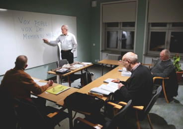 Man står framför whiteboard med text. Tre män är elever och deltar i undervisning.