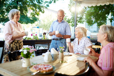 en samling seniorer sitter på ett utomhuscafé och fikar. det ser ut som en person berättar något roligt.