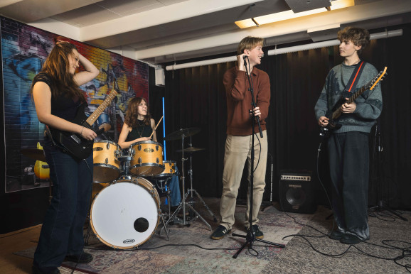 Fyra unga tjejer och killar med instrument i en replokal. De ser glada ut.