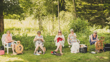 Fem kvinnor sitter i rad i naturen på trädgårdstolar, de arbetar med ulltråd, två spinner, en stickar.
