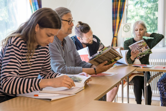 fyra personer sitt vid bord möblerat som skolsittning, alla läser ur samma bok