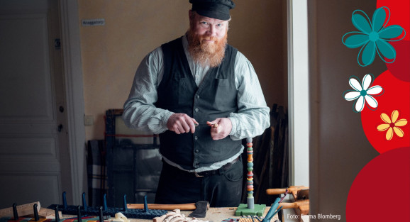 Frans Ekelin står i ett rum. Han är klädd i svart väst, blå skjorta och svart keps. I handen håller han en täljkniv samt ett bit trä. Framför honom syns ett bord med träföremål, bl.a. en hängare med krokar.