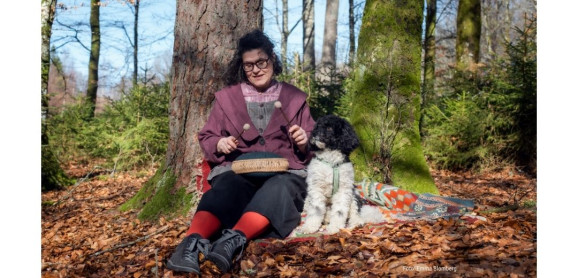 En kvinna sitter i en liten backe ute i naturen bland träden. Hon bär bland annat glasögon, en lila jacka, svarta byxor och röda strumpbyxor. I knät har hon ett litet instument som hon slår på med klubbor. Till höger om henne sitter en liten svart vit hund.