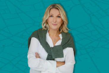 Annica Dahl tittar in i kameran med armarna i korv hon bär en vit skjorta och en grön kofta över axlarna. Bakgrunden är turkos