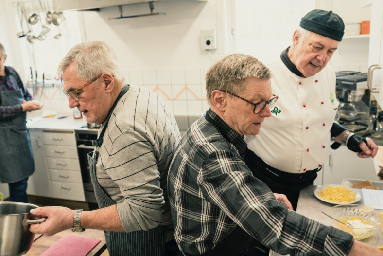 Två män som lagar mat under ledning av en kock i ett kök.