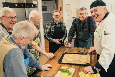 Grupbild från matlagningingcirkel med cirkelledare och cirkeldeltagare som står runt ett bord med rätter som förbereds.