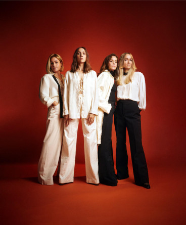 Pressbild på gruppen Sahara Hotnights som står i nästan enbart vita kläder mot en mörkt röd bakgrund.