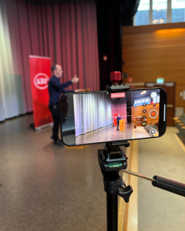 Tomas Eneroth syns från sidan genom en mobilkamera där han står vid ett podium på en scen och talar.