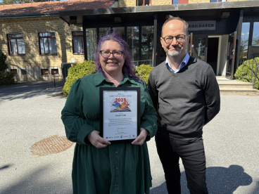 Eileen Falk, vinnare av Boken på arbetsplatsen 2025, håller i sitt diplom utomhus en solig försommardag. Brevid henne står Tomas Eneroth, ABFs förbundsordförande.