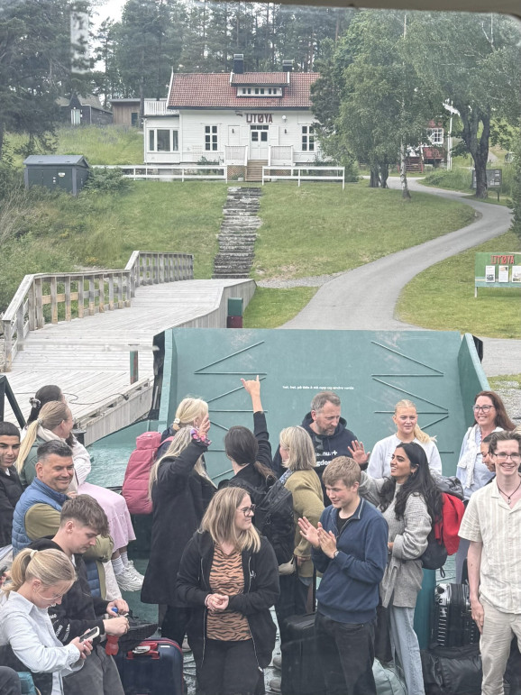 Många ungdomar står på färjan som lägger ut från Utøya. I bakgrunden ser man ett vitt hus med orden "Utøya" i röda bokstäver.