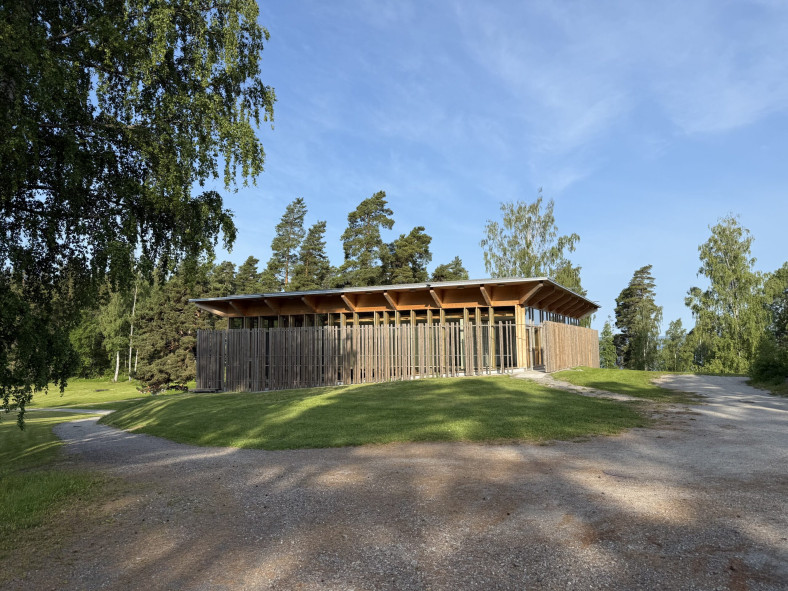 Hegnhuset som omgärdar den gamla samlingslokalen Kafébygget syns mot en blå himmel med grönska och träd i bakgrunden.