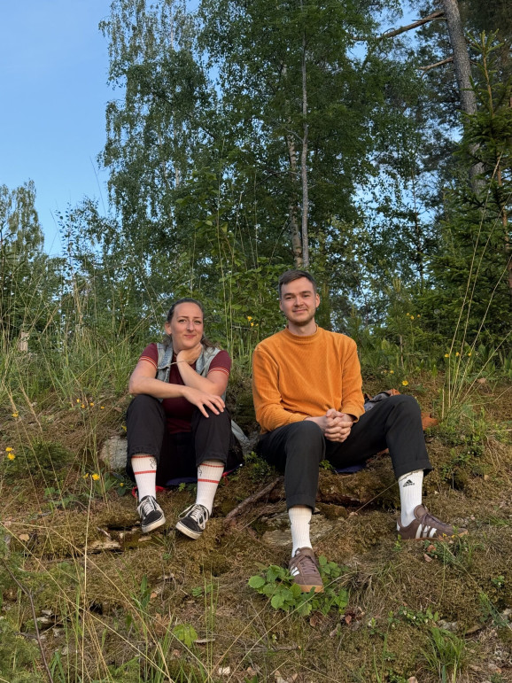 En kvinna och en man sitter brevid varandra utomhus i en slänt och tittar in i kameran.