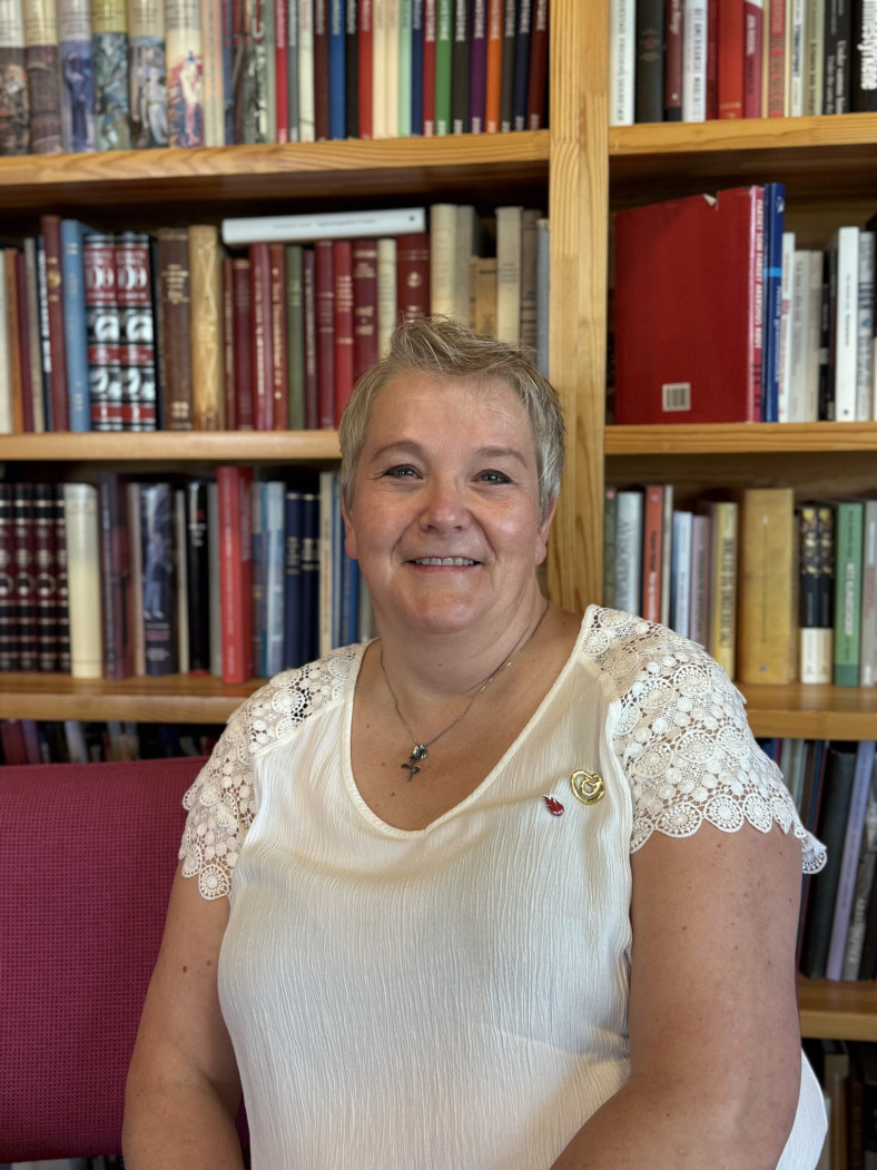 Monica Lund Schjetne sitter framför en bokhylla och tittar in i kameran med ett leende.