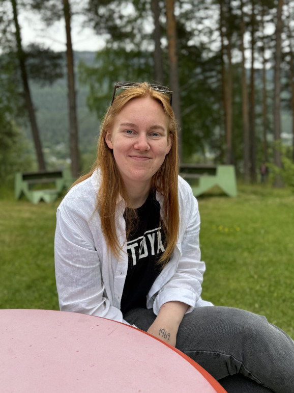 En närbild på en ung kvinna i vit skjorta sitter utomhus och tittar in i kameran med ett litet leende.