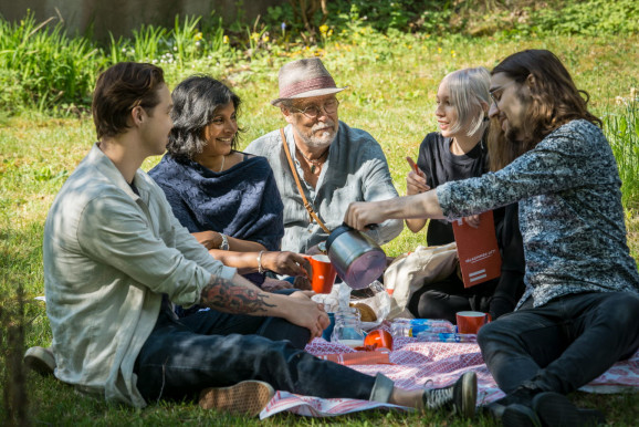 fem personer på picknickfilt dricker varm dryck och pratar