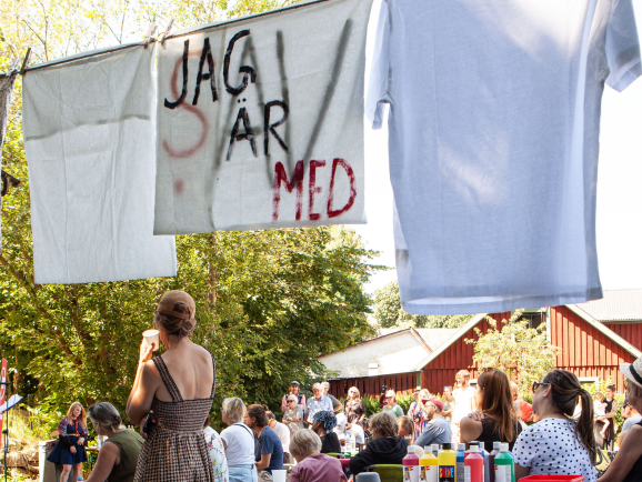 En bild som föreställer en samling människor som är utomhus vid en scen. Det står en skylt "Jag är med".
