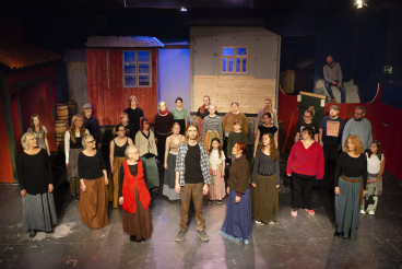 Skådespelare står på en scen, allas blickar är på en man i mitten av bilden