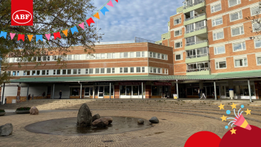 En öppen torgmiljö med en stenprydd vatteninstallation i förgrunden, omgiven av röda tegelbyggnader med butiker på bottenvåningen. ABF-logotypen syns i övre vänstra hörnet tillsammans med färgglada vimplar, och en partyikon med konfetti syns i nedre högra hörnet.