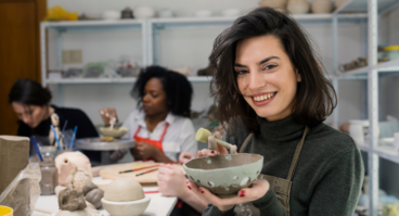 En leende kvinna håller upp en keramikskål i sina händer i en ljus keramikverkstad. I bakgrunden syns andra personer som arbetar med keramik vid drejskivor och hyllor fyllda med keramikföremål.