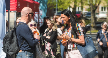 En grupp människor minglar utomhus. I förgrunden syns en kvinna i en randig blus som ler och talar med en man som bär solglasögon och en ryggsäck. De håller båda i dryckesflaskor.