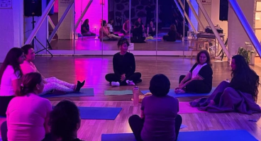 Grupp av personer sitter i cirkel på yogamattor i ett rum med trägolv och spegelvägg. Rummet är upplyst med lila och rosa belysning, vilket skapar en lugn och kreativ atmosfär.