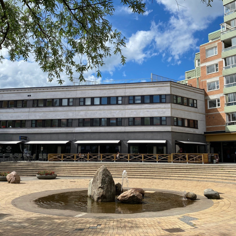 Fasad mot Poseidons torg med en låg byggnad i flera våningar, uteservering och en rund fontän med stenar i förgrunden.