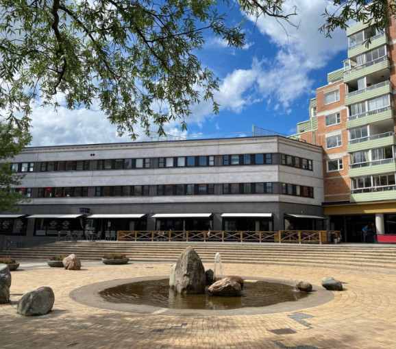Fasad mot Poseidons torg med en låg byggnad i flera våningar, uteservering och en rund fontän med stenar i förgrunden.