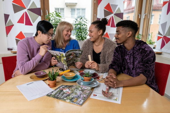 fyra personer pratar vid ett bord och tittar ner i en tidning på bordet finns planterade växter