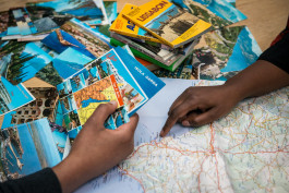 två händer syns på bilden, den ena tittar på vykort och den andra pekar på en ort i Spanien på en karta