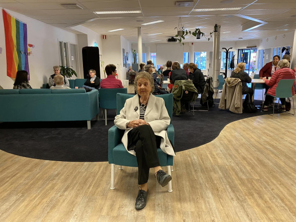 Bild en folksamling som sitter i ett caé och lyssnar på en föreläsning. En äldre dam sitter ensam i mitten vänd mot kameran